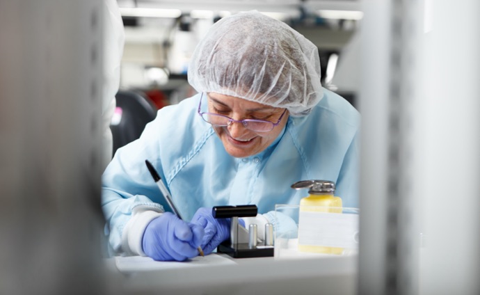 Smiling laboratory worker in medical gear writing notes next to lab equipment.